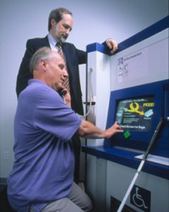 Dr. Gregg Vanderheiden standing by to assist a user sitting in front of a kiosk screen.