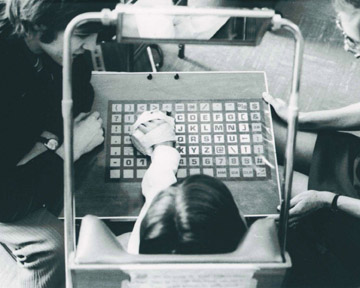 An overhead shot of a child, hand outstretched and taped to a handpiece on the surface of a communication board with a grid of letters, numbers, and symbols; a man on their left and a woman on their right.