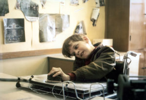 A boy sitting in a wheelchair moves a handpiece across the top of an auto monitoring communication board