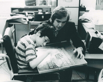 An overhead shot of a child, hand outstretched and taped to a handpiece on the surface of a communication board with a grid of letters, numbers, and symbols