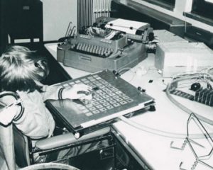 Boy in wheelchair using sliding hand-piece to point to letters on an aluminum communication board wired to a typewriter.