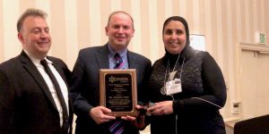 Dr. Lazar stands between two meeting attendees, holding an award plaque.